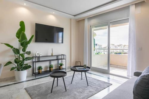 Modern living room in Marbella apartment with flat-screen TV, black furniture, potted plant, and balcony overlooking Puerto