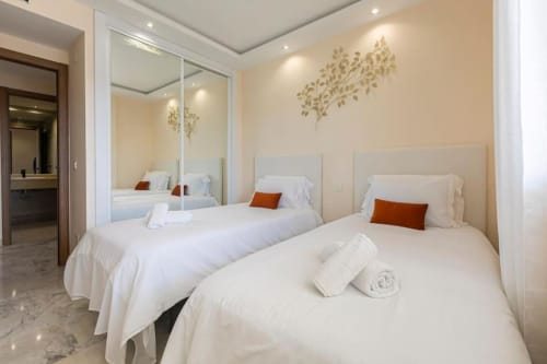 Two twin beds with white linens and orange pillows in a bright Marbella apartment at Marina Banús, featuring mirrored