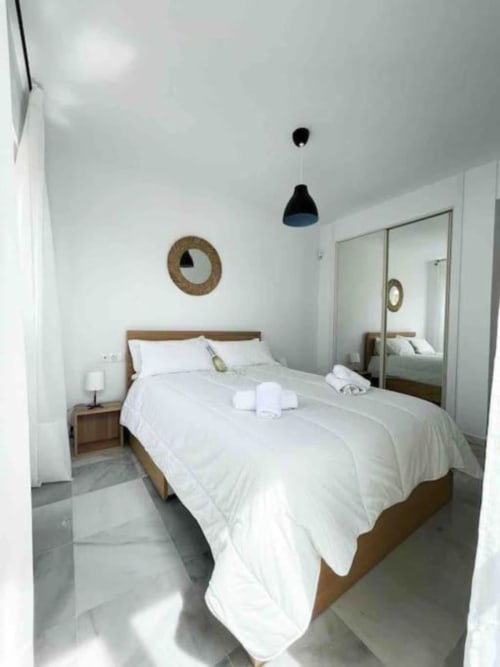 Modern bedroom in Marbella apartment with wooden bed, white linens, pendant light, and mirrored wardrobe