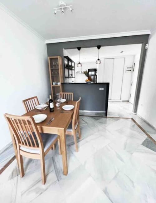 Modern dining area with wooden table and chairs in Marbella apartment, open kitchen with gray counter and pendant lights