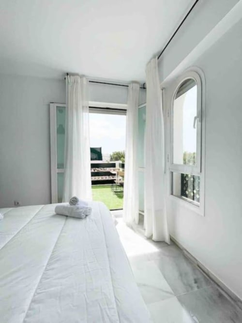 Bright Marbella apartment bedroom with white bed, garden view through open French doors, and wooden balcony overlooking green