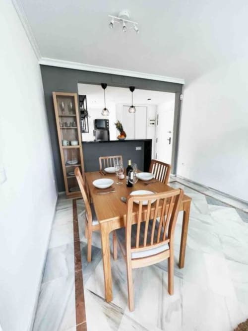 Modern dining area with wooden table and chairs, open kitchen with dark counters, pendant lights, and white walls in Marbella