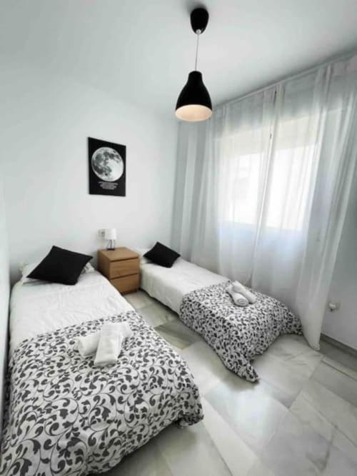 Twin bedroom in Marbella apartment with patterned bedding, wooden nightstand, black pendant light, and moon wall art.