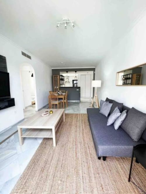 Modern open-plan living room and kitchen in Marbella apartment with gray sofa, dining area, and garden views