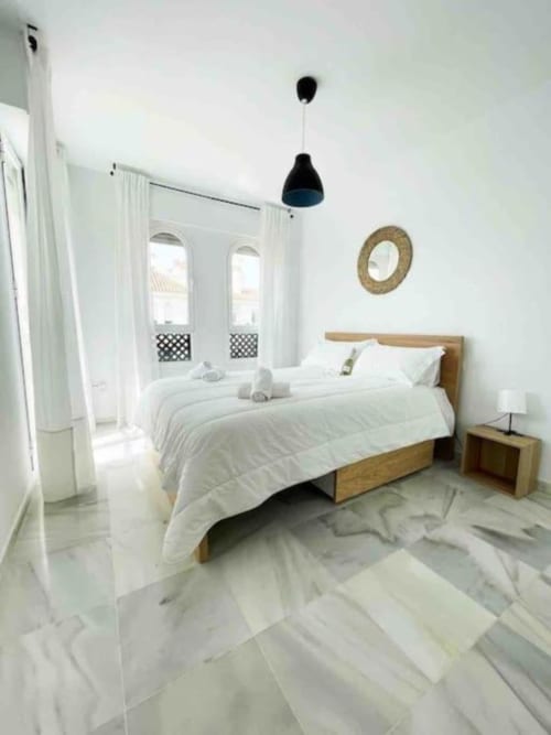 Bright modern bedroom in Marbella apartment with wooden bed, white linens, marble floors, black pendant light, and balcony