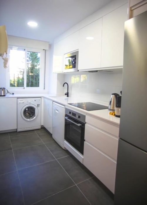 Modern white kitchen with stainless steel appliances, black tile flooring, and washing machine in Marbella apartment