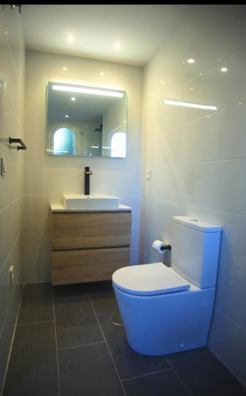 Modern bathroom in Marbella apartment with blue toilet, white vessel sink, wooden vanity, and illuminated mirror.