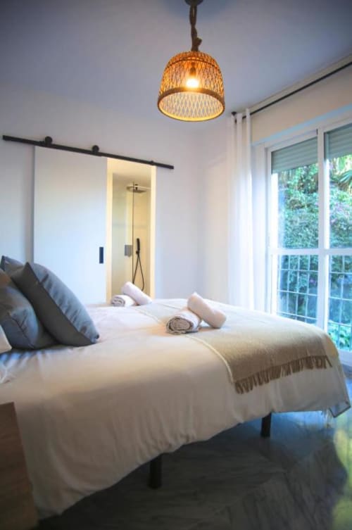 Modern bedroom in Marbella apartment with comfortable bed, woven pendant light, ensuite bathroom, and terrace doors