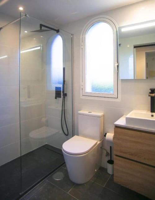 Modern bathroom in Marbella apartment with glass shower, white toilet, wooden vanity, and arched window overlooking pool area