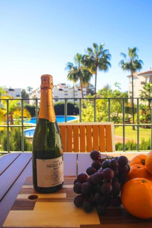 Champagne bottle with grapes and oranges on terrace at Marbella apartment with pool view and palm trees