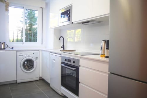 Modern kitchen in Marbella apartment with white cabinetry, stainless steel appliances, washing machine, and natural light