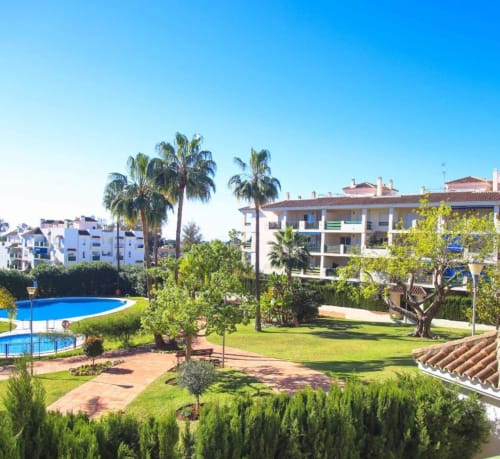 Marbella apartment complex with pool view, palm trees, lush gardens, and modern Mediterranean architecture in Nueva Andalucía