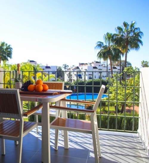 Sunny terrace balcony with dining table, pool view, palm trees, and white apartments in Marbella, Spain