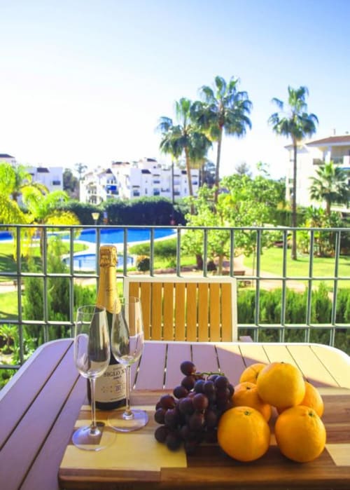 Elegant balcony at Marbella apartment with champagne, fresh fruit, and pool views overlooking lush gardens and palm trees