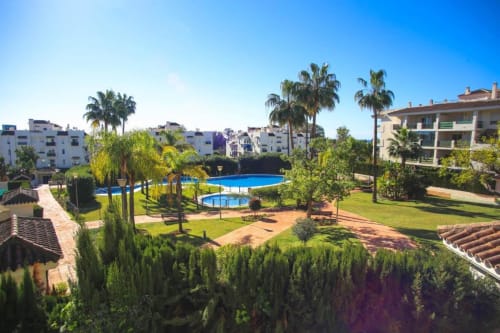 Sunny pool area at Marbella Home Rentals with palm trees, loungers, and residential buildings in Nueva Andalucía, Spain