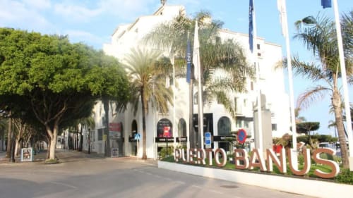 Puerto Banús promenade in Marbella with white building, palm trees, and sea view - First Line Apartment location