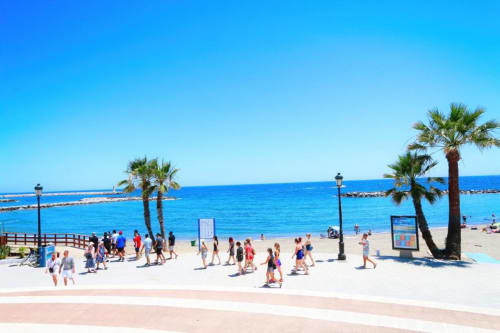 Paseo marítimo soleado de Marbella con palmeras, playa arenosa, aguas azules del Mediterráneo y bañistas cerca del Puerto