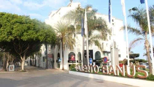 Letrero de entrada a Puerto Banús con edificio blanco, palmeras y banderas en Marbella, España