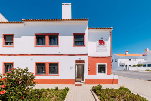 Exterior do hotel Casa da Praia com paredes brancas e laranja, telhado de barro e plantas floridas em Zambujeira do Mar