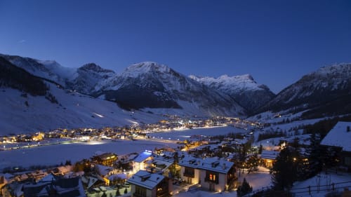 Valle alpina innevata con villaggio illuminato e lago al tramonto, Livigno Italia