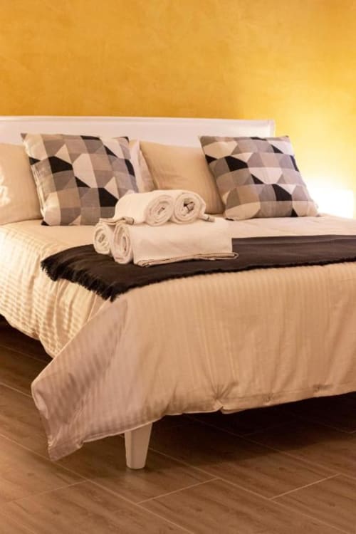 Letto elegantemente arredato a Dimore Verona con cuscini a motivi geometrici, biancheria color crema, asciugamani arrotolati e parete dai toni dorati