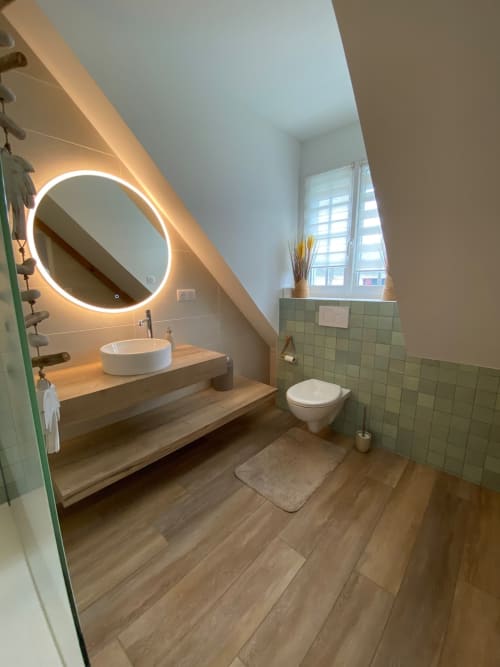 Salle de bain moderne avec miroir circulaire illuminé, lavabo blanc vasque, meuble en bois et accents de carrelage vert chez Lady Jule