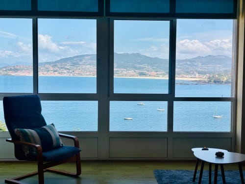 Casa de piedra costera en Baiona con vistas al mar, sillón azul, veleros y montañas al fondo