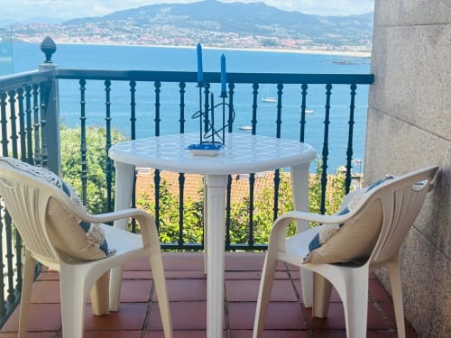 Terraza frente al mar con mesa y sillas blancas con vista a la bahía de Baiona, montañas y pueblo costero en España