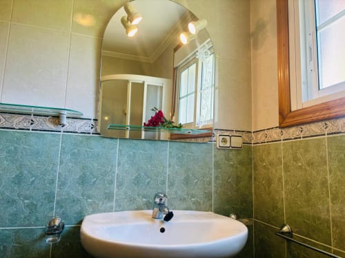 Baño moderno de la casita Xoaniña en Baiona con azulejos verde azulado, lavabo blanco redondo, espejo arqueado e iluminación cálida