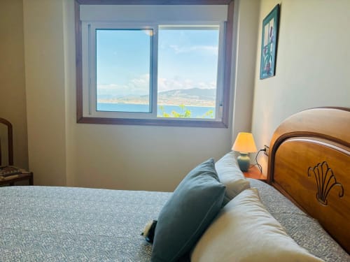 Dormitorio con vistas al mar de la costa de Baiona, cama de madera, lámpara de mesilla y ventana con vistas a las montañas y la bahía.