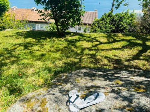 Sandalias blancas en terreno rocoso con jardín exuberante, cabaña con tejado de terracota y vista al mar en Baiona