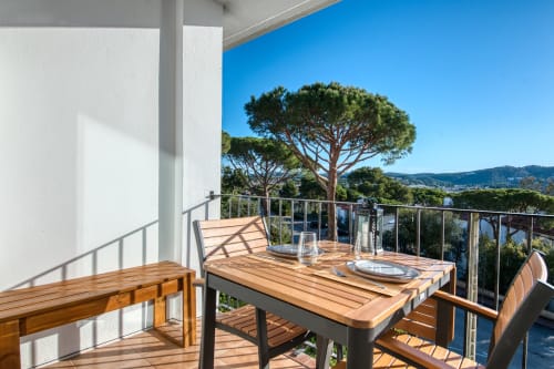 Sunny apartment terrace in Palafrugell with dining table, wooden chairs, sea views, and umbrella pine tree against clear blue