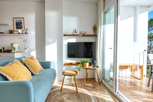 Bright modern apartment living room with blue sofa, sea views through glass doors, TV, wooden accent tables, and access to