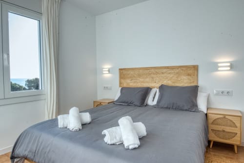 Modern bedroom with sea views, wooden headboard, gray bedding, white pillows, and natural light in Palafrugell apartment