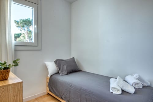 Bright bedroom with gray bed, white pillows, rolled towels, wooden nightstand with potted plant, and window overlooking tree