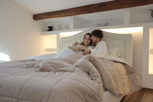 Coppia che si rilassa nella luminosa camera moderna dell'appartamento Enjoy House Bracciano con illuminazione ambientale e soffitto con travi in legno