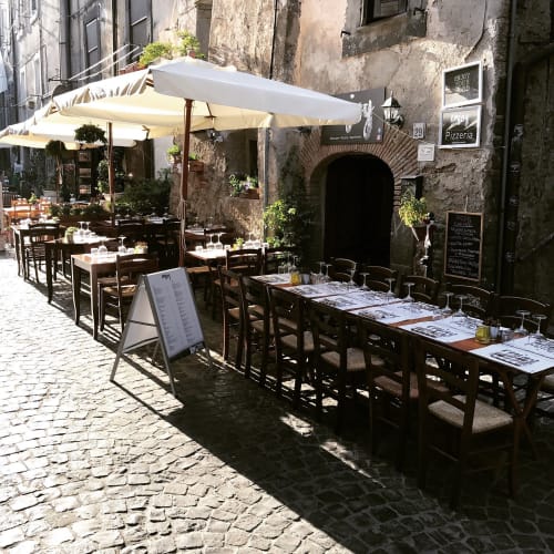Patio di una pizzeria all'aperto a Bracciano con ombrelloni bianchi, tavoli in legno e antichi edifici in pietra
