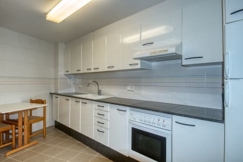 Modern kitchen in Palafrugell apartment with white cabinets, dark granite countertops, white appliances, and dining table.