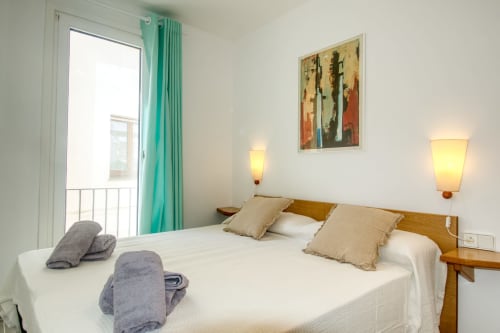 Bright bedroom with white bedding, wooden headboard, turquoise curtains, balcony door, and modern wall sconces at Finques