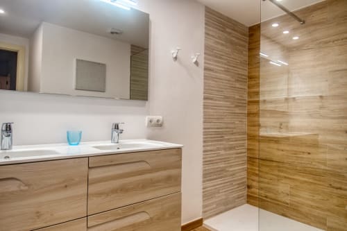 Modern bathroom in Palafrugell apartment with wood-grain tiles, white sink, large mirror, and glass shower enclosure