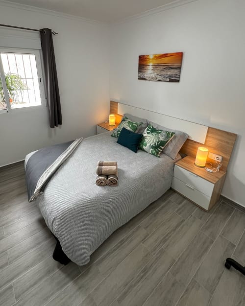 Dormitorio moderno en Casa Torrens con arte de atardecer costero, vista al océano, mesitas de noche de madera y toallas enrolladas.