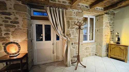 Entrée de cottage en Corrèze avec murs de pierre, rideaux beige, mobilier bois et éclairage chaleureux