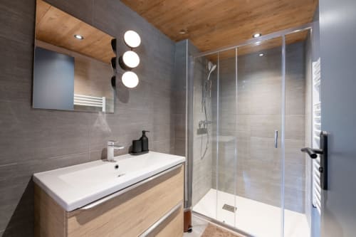 Salle de bain moderne du chalet Domaine de l'Epigny avec vasque suspendue, miroir illuminé, douche à l'italienne et plafond en bois.