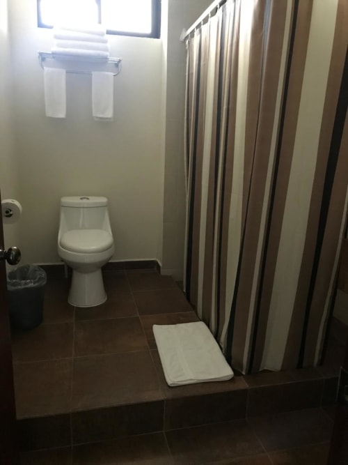 Baño del Hotel El Candelero en Yuriria con inodoro, cortina de ducha, toallero y piso de azulejos oscuros