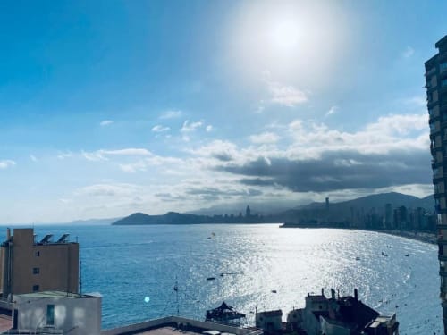 Sunny Benidorm beach view from apartment balcony with Mediterranean Sea, distant mountains, and coastal cityscape.