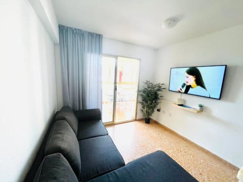 Modern living room in Mariscal Apartments, Benidorm with dark sofa, flat-screen TV, balcony with city views, and light wood