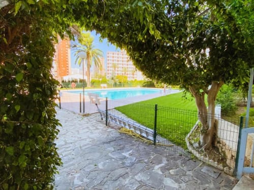 Garden pathway through lush greenery leading to resort pool with palm trees and apartment buildings at Tads Holiday Lets,
