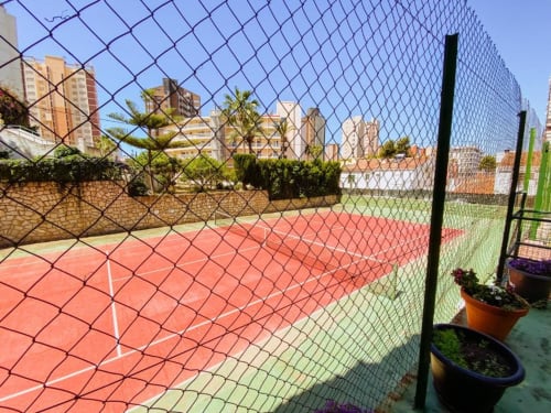 Tennis court with red clay surface at Zafiro Apartments in Benidorm, surrounded by chainlink fencing and city buildings