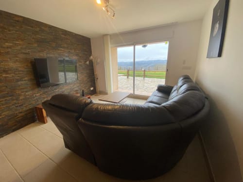 Salon de villa moderne avec canapé noir courbé, foyer en brique, TV et vue sur les montagnes par portes vitrées coulissantes au
