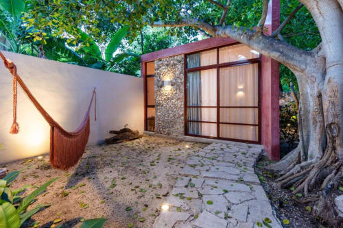 Patio de la posada Casa Tuch en Mérida con estructura roja moderna, detalles en piedra, hamaca y árbol frondoso